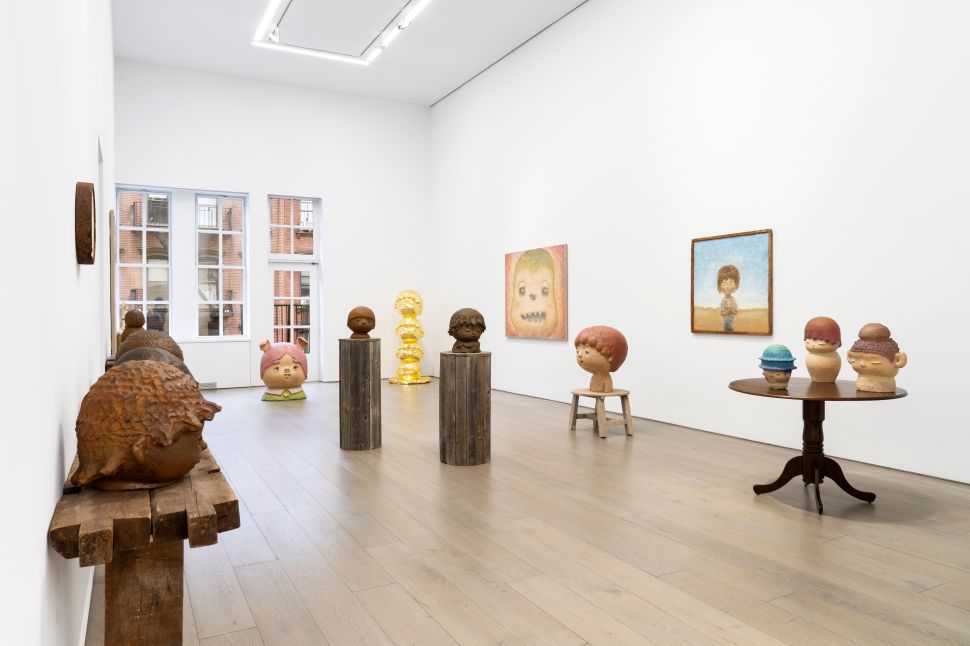 Installation view of a contemporary art exhibition showcasing large, playful ceramic sculptures of humanlike figures on wooden pedestals. Several smaller figures are displayed on a wooden table. The surrounding wall features colorful paintings, including portraits of figures with exaggerated features, adding to the whimsical atmosphere of the space.