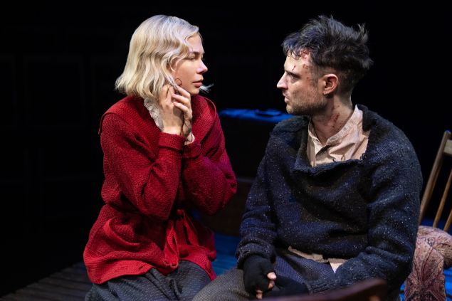 An image shows a woman in a red sweater sitting close to a man with visible cuts on his face, both turned toward each other in an intense, quiet exchange on a dark stage.
