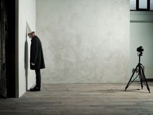 A man wearing a white conical hat stands with his forehead pressed against a blank wall while a video camera on a tripod records him from across an empty room, creating a scene of deliberate absurdity characteristic of Erwin Olaf’s April Fool images.