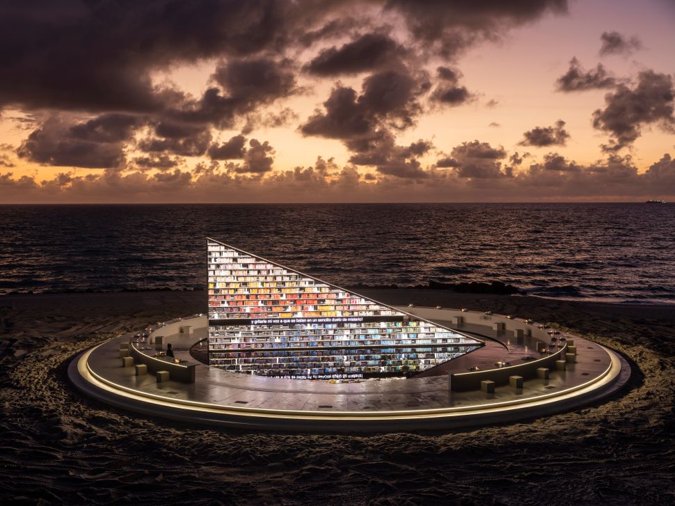 A large triangular mirrored structure filled with colorful book spines stands on the sand at the edge of the ocean at sunset, surrounded by a circular platform glowing with soft light.