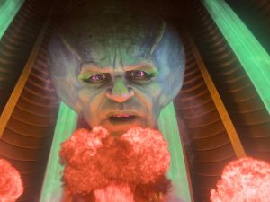 A giant projected head with greenish skin and glowing eyes looms over fiery bursts of red smoke, representing the exaggerated Wizard figure in Sphere’s reimagined version of the film.