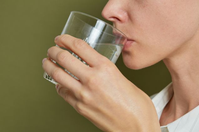 A woman's hands holding a glass of water, seen from the side against a green background.