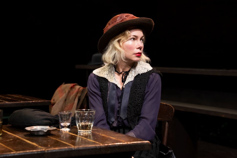 An image shows a woman wearing a hat and dark vest seated alone at a wooden table with glasses, looking off to the side under focused stage lighting in an otherwise dark space.