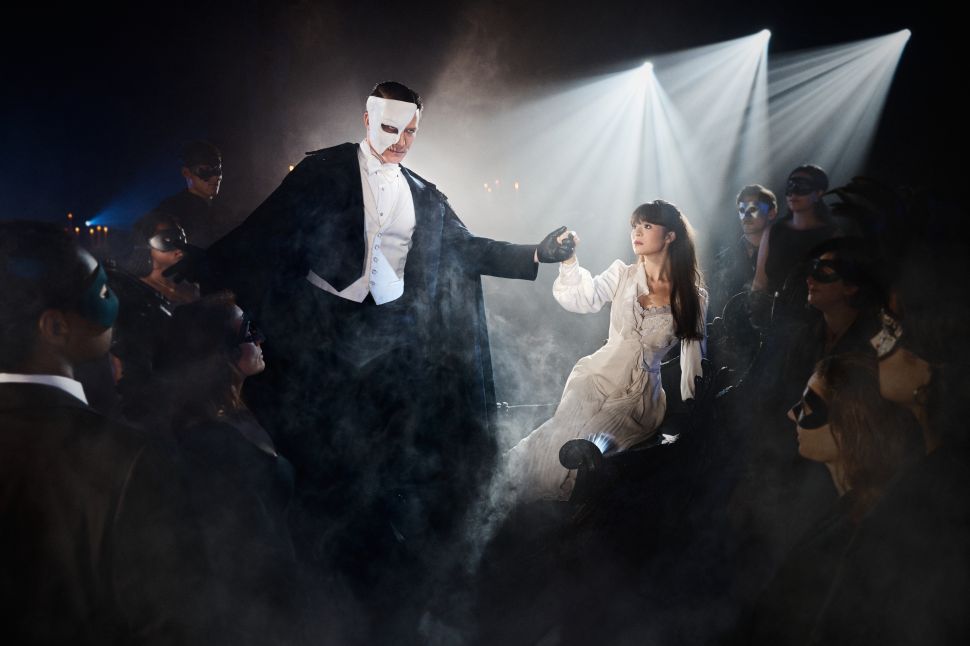 An actor dressed as the Phantom in a half-mask holds the hand of an actress as Christine while surrounded by masked audience members in a smoky immersive theater scene.