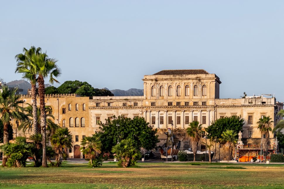 A wide view of Palazzo De Seta in Palermo, with its neoclassical façade framed by palm trees and an open lawn in the foreground, set against distant mountain silhouettes under a clear sky.