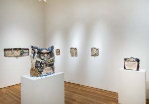 A gallery installation view shows multiple ceramic works arranged on the walls and two large ceramic sculptures on white pedestals, all illuminated by overhead spotlights on wooden floors. If you'd prefer these written in your Observer alt-text style (more concise, more atmosphe