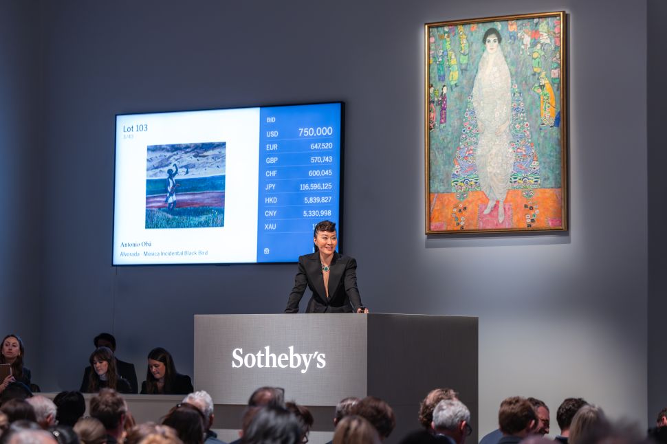 A Sotheby’s auctioneer in a black suit stands in front of a crowded room with Gustav Klimt’s portrait of a woman hanging beside a bidding screen showing Antonio Obá’s Alvorada—Musica Incidental Black Bird.