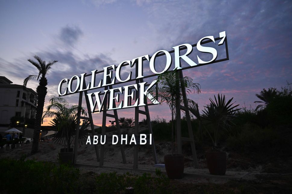 A large illuminated sign reading "Collectors’ Week Abu Dhabi" stands against a purple sunset sky, surrounded by palm trees and greenery.