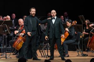 An image of two male opera singers performing on stage in formal tuxedos with the orchestra in the background, as they sing passionately into microphones, with an orchestra seated behind them, including a cellist, amidst a dark background highlighting the singers' dramatic expression.