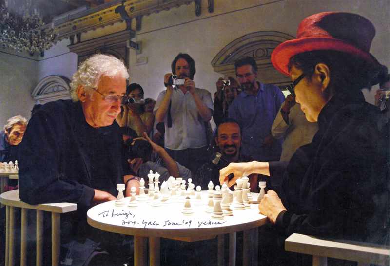Luigi Bonotto plays a white chess set with Yoko Ono, who wears a red hat, as photographers gather around them in a crowded room.