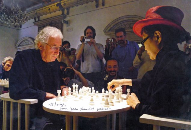 Luigi Bonotto plays a white chess set with Yoko Ono, who wears a red hat, as photographers gather around them in a crowded room.